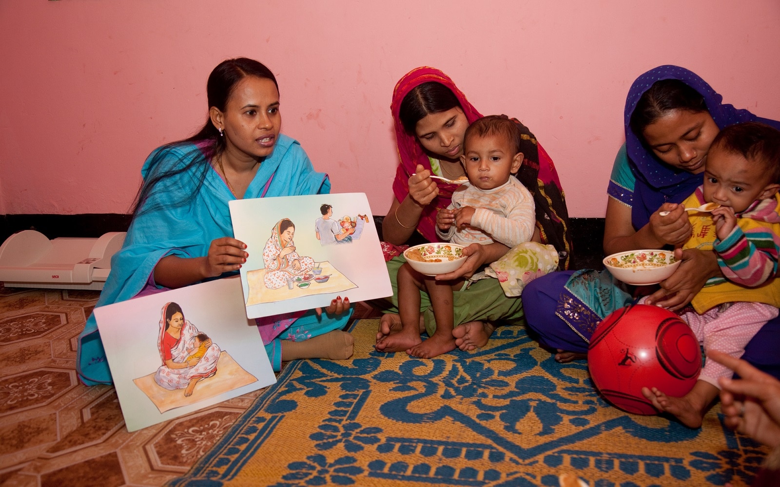 Children covered under ICDDR,B's Malnutrition-Enteric Disease (Mal-ED) Study receive food supplements while their mothers receive nutrition information from Field Research Assistant, Parveen Sultana Mita (with picture cards), at a Community Nutrition Centre in the Mirpur locality. 